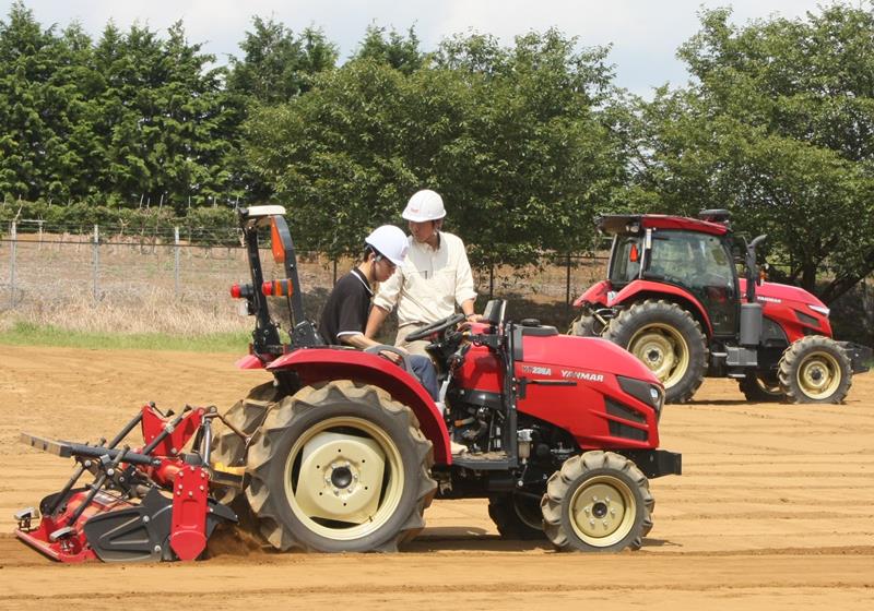 直進アシスト付きトラクターの運転を体験する学生。写真奥は自動運転トラクター＝千葉市緑区