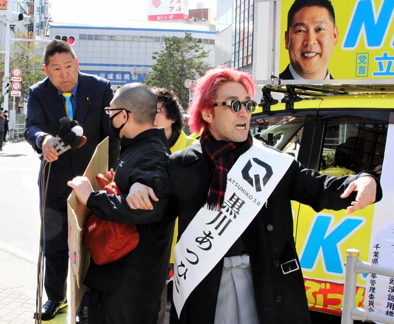 JR船橋駅前での第一声でプラカードを掲げる聴衆にマイクを向ける立花氏(左)と、聴衆を誘導する黒川氏(右)=2月27日、船橋市